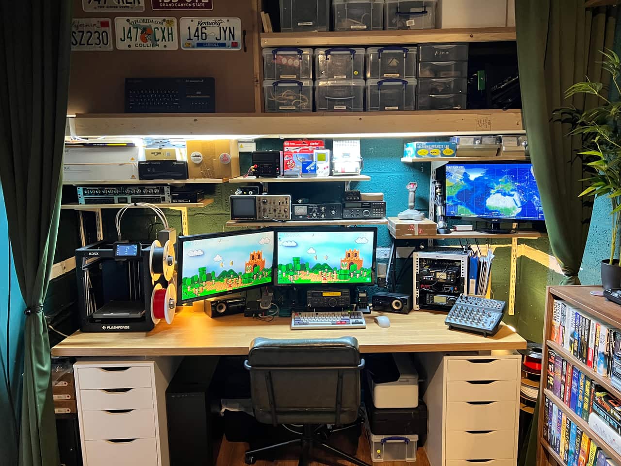 A workbench glowing in a dark room: monitors, a 3D printer, electronics and shelves of parts.